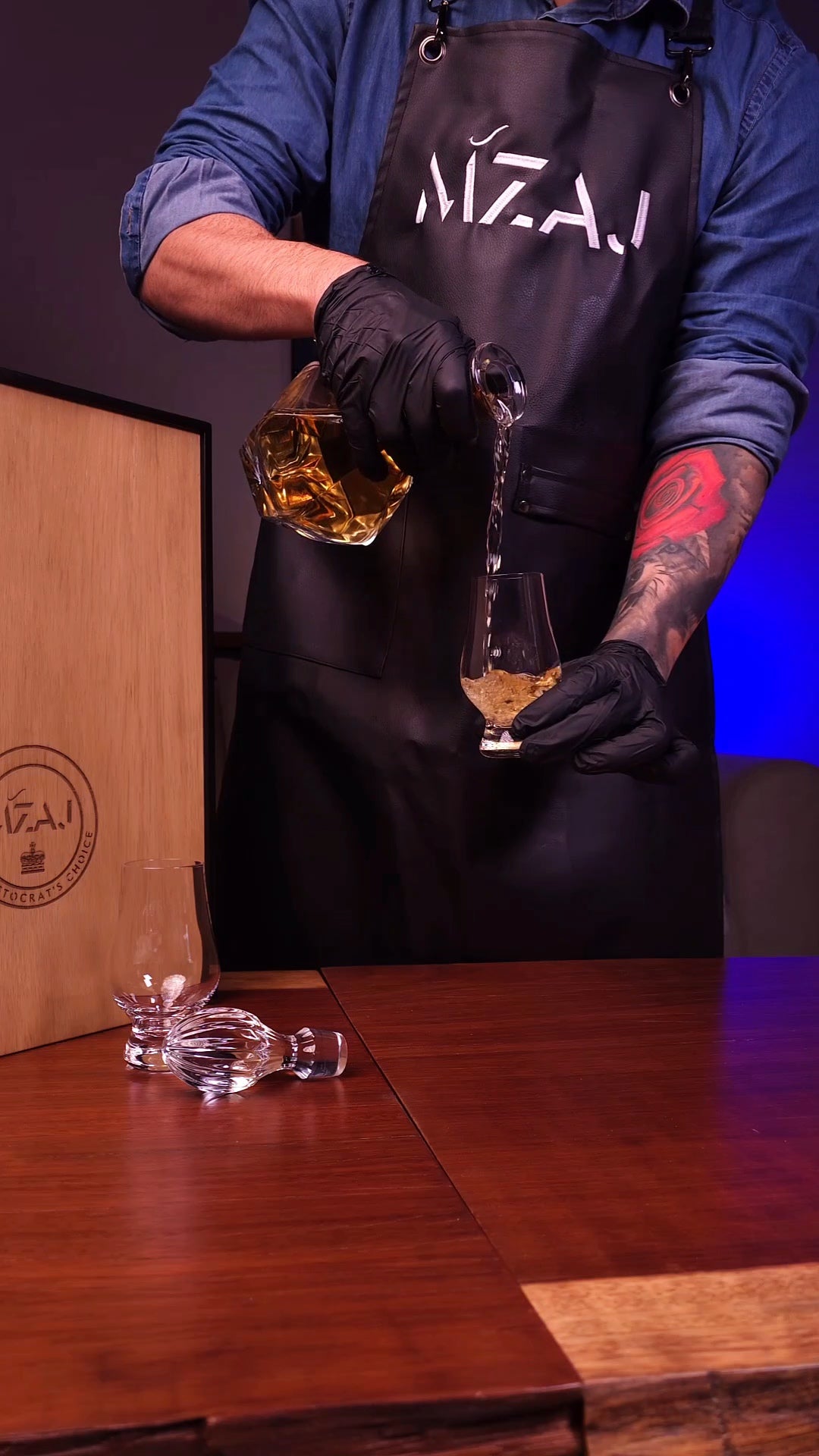 Person in a Mzaj branded apron pouring a drink into a glass on a wooden table.